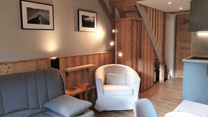 Una stanza accogliente con un divano grigio, una poltrona bianca, travi in legno e una scala che conduce a una zona cucina.