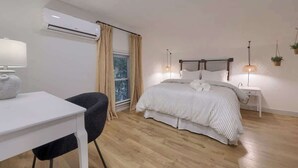Un dormitorio con cama, mesitas de noche, escritorio, una silla, unidad de aire acondicionado y ventana con cortinas.