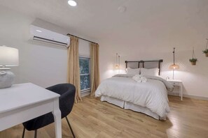 Un dormitorio con cama, mesitas de noche, escritorio, una silla, unidad de aire acondicionado y ventana con cortinas.