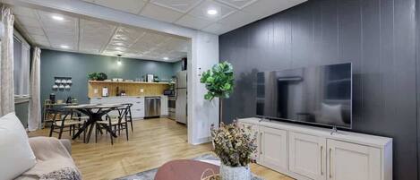A modern living room with a dining area, a kitchen in the background, and a large flat-screen TV mounted on the wall.