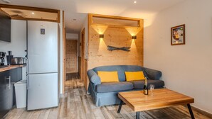A compact living space with a kitchenette, a sofa with yellow cushions, a wooden table with a bottle and glass, and a wooden wall with a light fixture. - Cosy T2 apartment with terrace 5 min from Avoriaz cable car (Morzine)