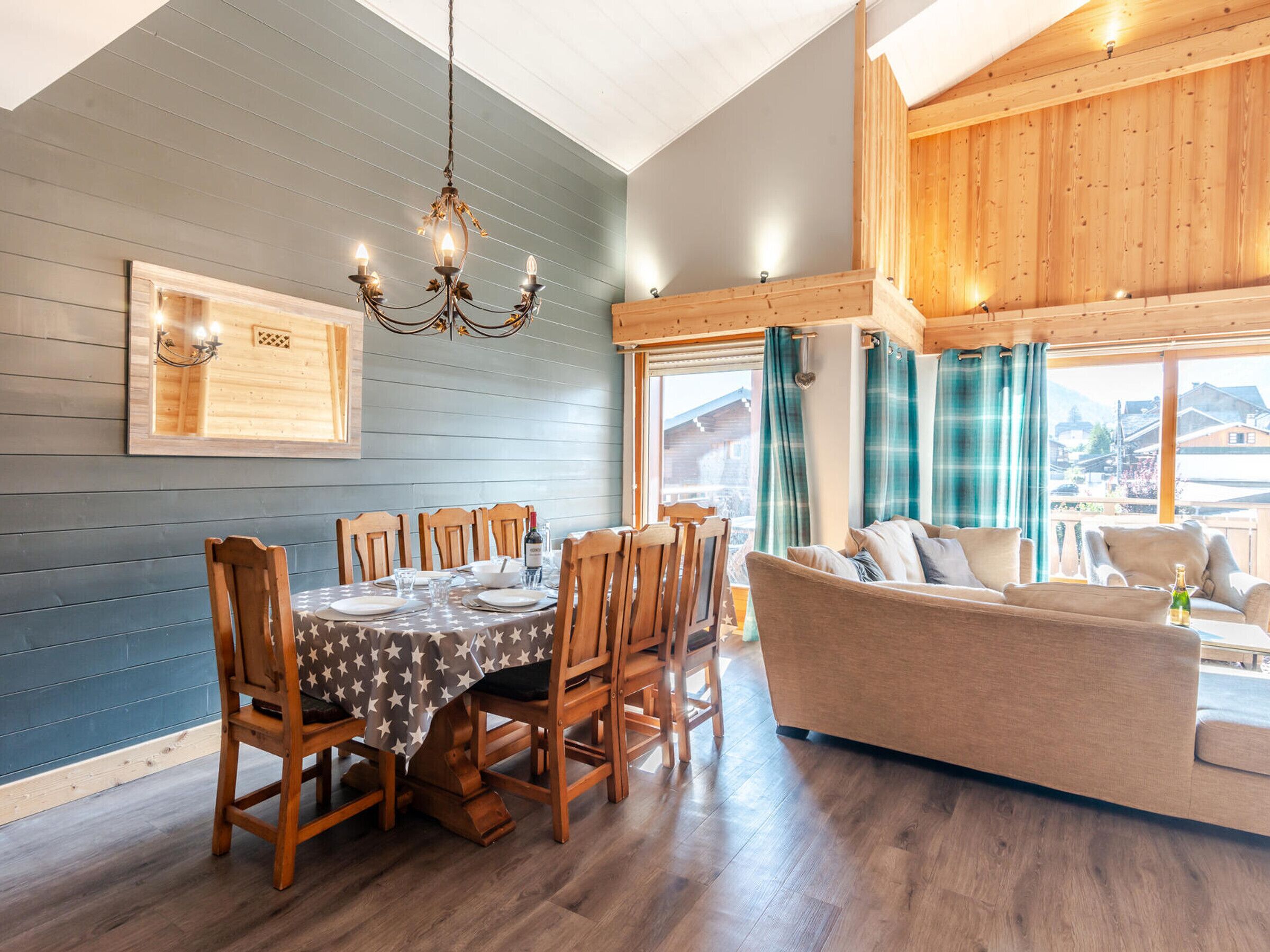 Un coin repas avec une table dressée pour six personnes, des chaises en bois, un lustre et une vue sur l’extérieur grâce à de grandes fenêtres.