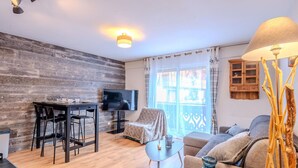 A modern living room with a wooden accent wall, a flat-screen TV, a dining area with chairs, a sofa, and a lamp. - Renovated studio-cabin for 4, central Morzine, near slopes (Morzine)