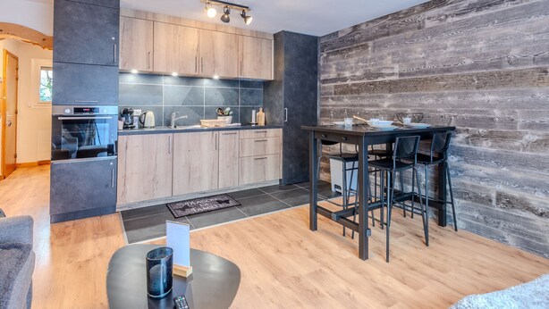 A modern kitchen with dark cabinets, a wooden dining table, and a rustic wall. - Renovated studio-cabin for 4, central Morzine, near slopes (Morzine)