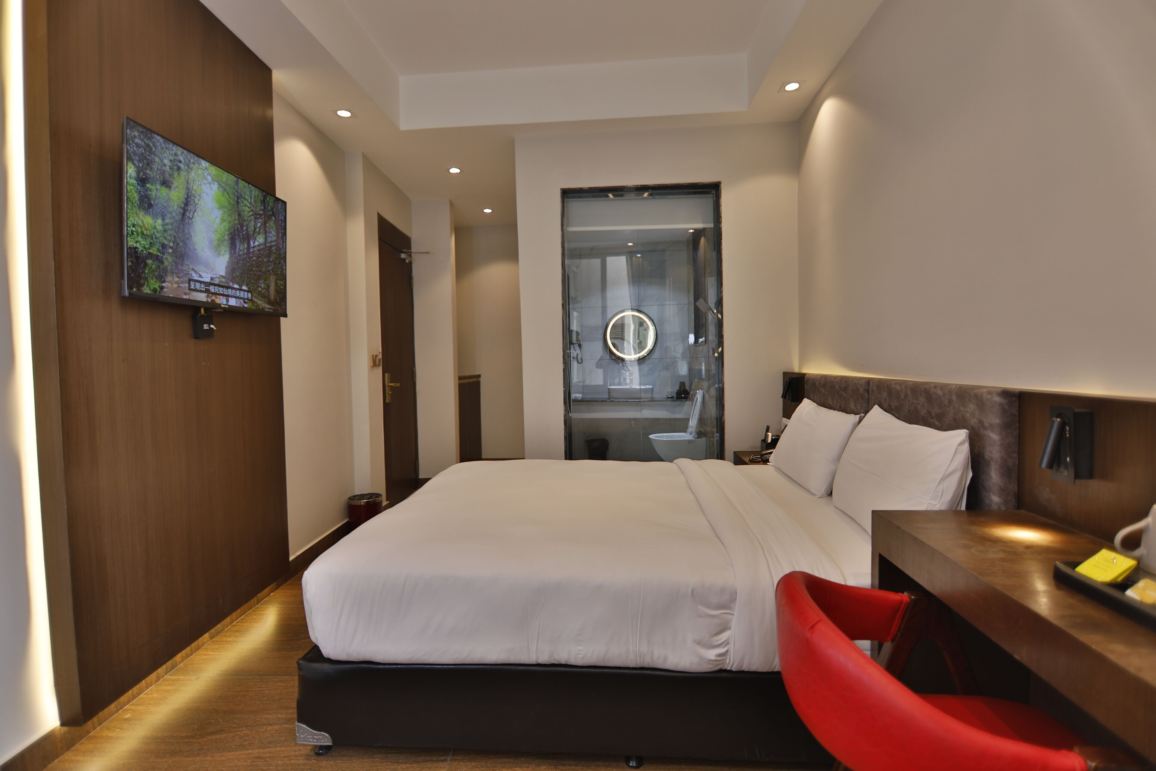 A modern hotel room with a large bed, a TV mounted on the wall, a desk with a chair, and a bathroom visible through a glass door.