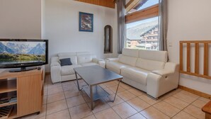 Una sala de estar moderna con un sofá de cuero blanco, una mesita de centro y un televisor sobre un mueble de madera. Un ventanal ofrece vistas a montañas y edificios.