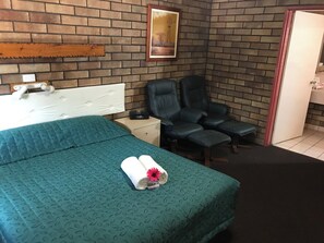 A hotel room with a bed, a chair, a nightstand, a picture on the wall, and a door. - Miles Outback Motel (Miles)