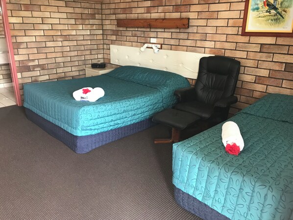 A room with two beds, a chair, a brick wall, and a framed picture.