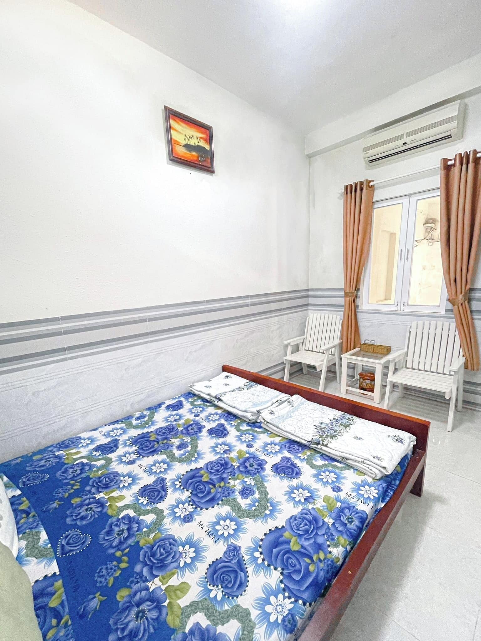 Un dormitorio con cama, una mesita, dos sillas, una ventana con cortinas, aire acondicionado y un cuadro en la pared.