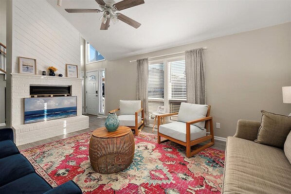 A living room with a fireplace, two chairs, a sofa, a ceiling fan, and a large window with blinds.