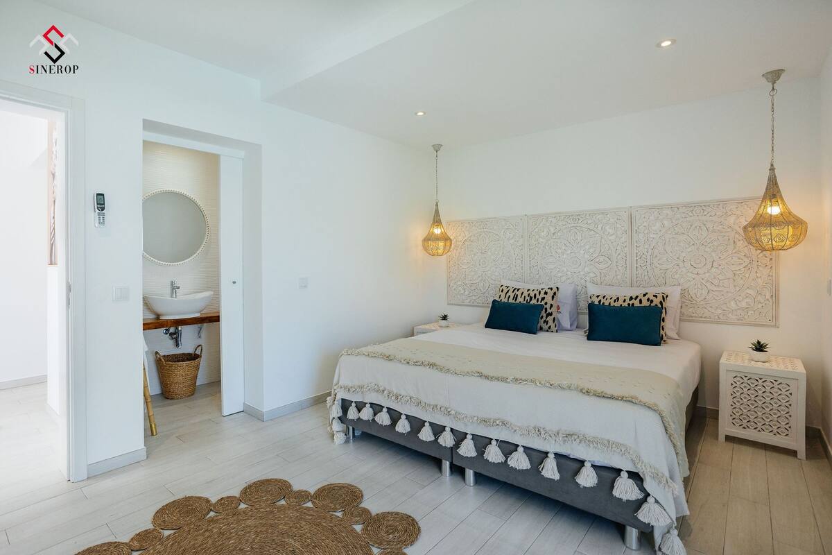 Une chambre à coucher comprenant un lit, des tables de chevet, un luminaire suspendu, un miroir fixé au mur et une porte donnant accès à une salle de bain.