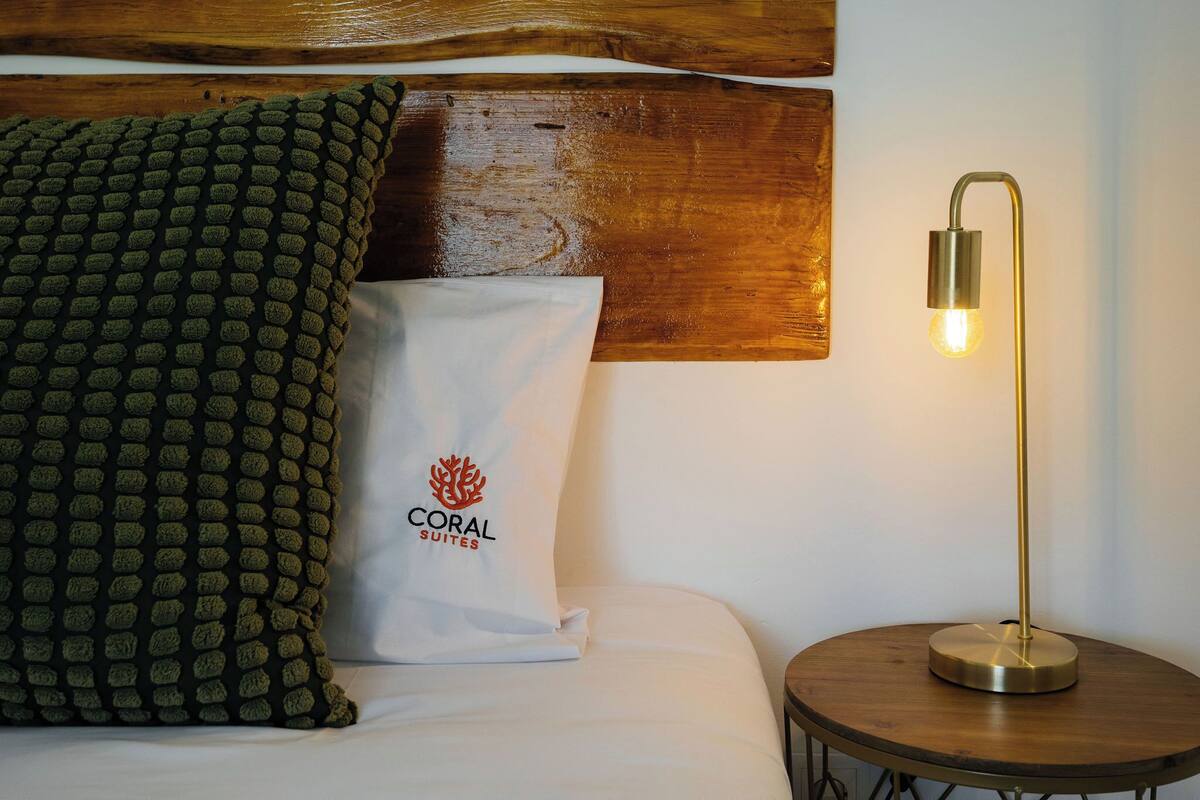 Un lit bien fait, agrémenté d’un coussin vert à texture et d’un coussin blanc orné d’un logo, une table d’appoint ronde en bois avec une lampe, et une tête de lit en bois.
