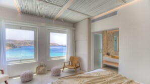 A modern bedroom with a large bed, a wooden chair, and a view of the sea through large windows. - Icon Mykonos (Mykonos)