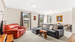 A spacious living room with a red leather sofa, a black leather sectional, and a wooden coffee table. Large windows provide natural light and views of the outdoors. - The Plover Escape Phillip Island (Cowes)