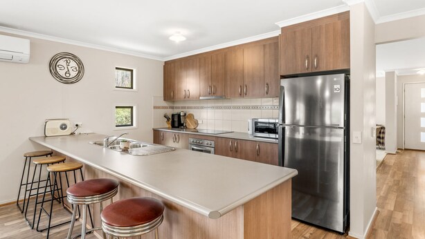A modern kitchen with wooden cabinets, stainless steel appliances, a large island with stools, and a view into the adjacent living area. - The Plover Escape Phillip Island (Cowes)
