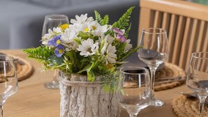 Una mesa redonda de madera dispuesta para comer, con un centro de mesa rústico de flores en un recipiente texturizado, rodeada de vasos de vidrio y manteles individuales tejidos.