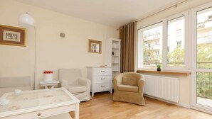 A modern living room with a glass coffee table, white furniture, a beige armchair, a white cabinet, and a large window with a view of buildings.