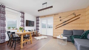 A modern living room with a wooden wall, a grey sofa, a dining area with a wooden table and chairs, and ski equipment mounted on the wall. - Cozy Apartment | With Mountain View and Balcony (Koscielisko)