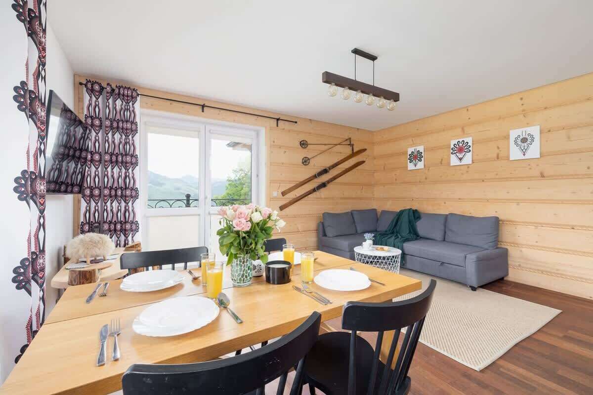 A dining area with a wooden table set for two, a grey sofa, and a view of the outdoors.