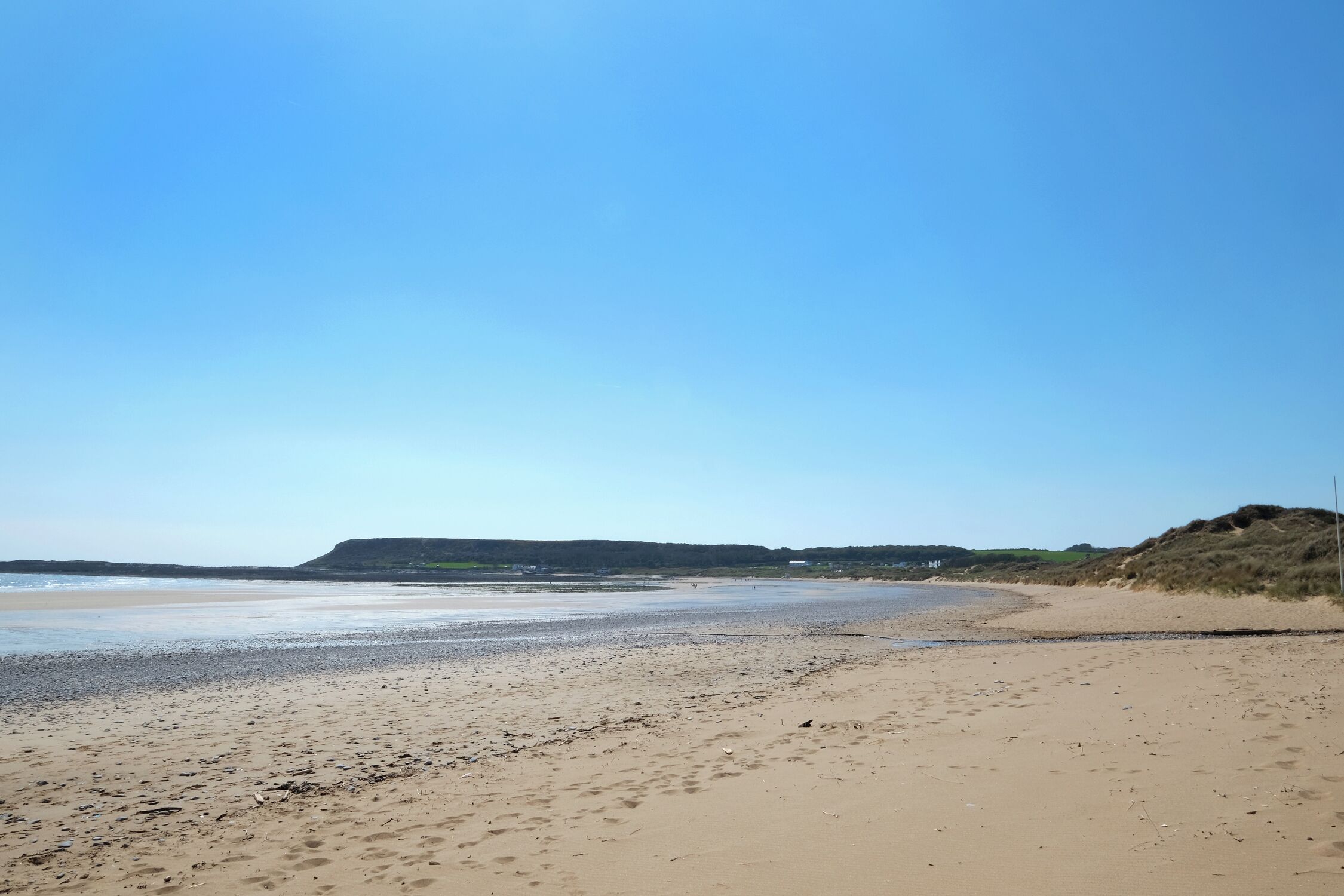 Une vaste plage de sable fin, avec des vagues douces, un ciel dégagé et des collines à l’horizon.