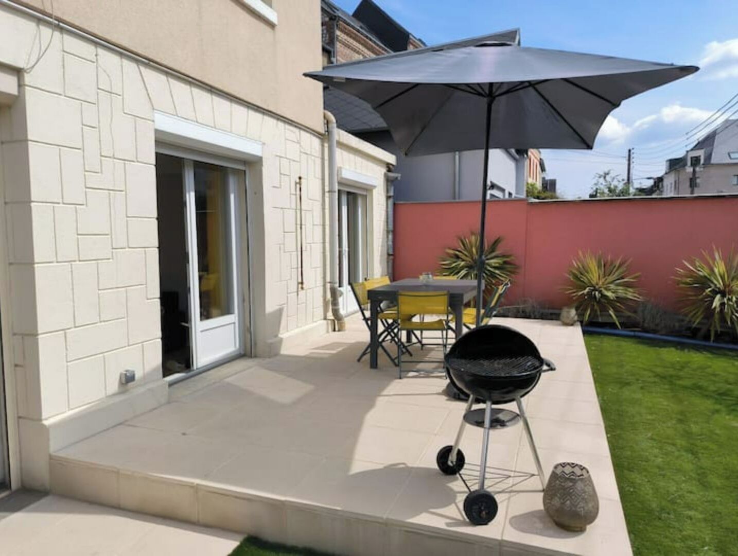 Un espace extérieur aménagé en patio, avec un barbecue, une table entourée de chaises et un grand parasol pour créer de l’ombre.