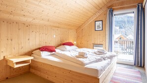 Un dormitorio de cabaña de madera con cama, almohadas, una mesita de noche y una ventana con cortinas.