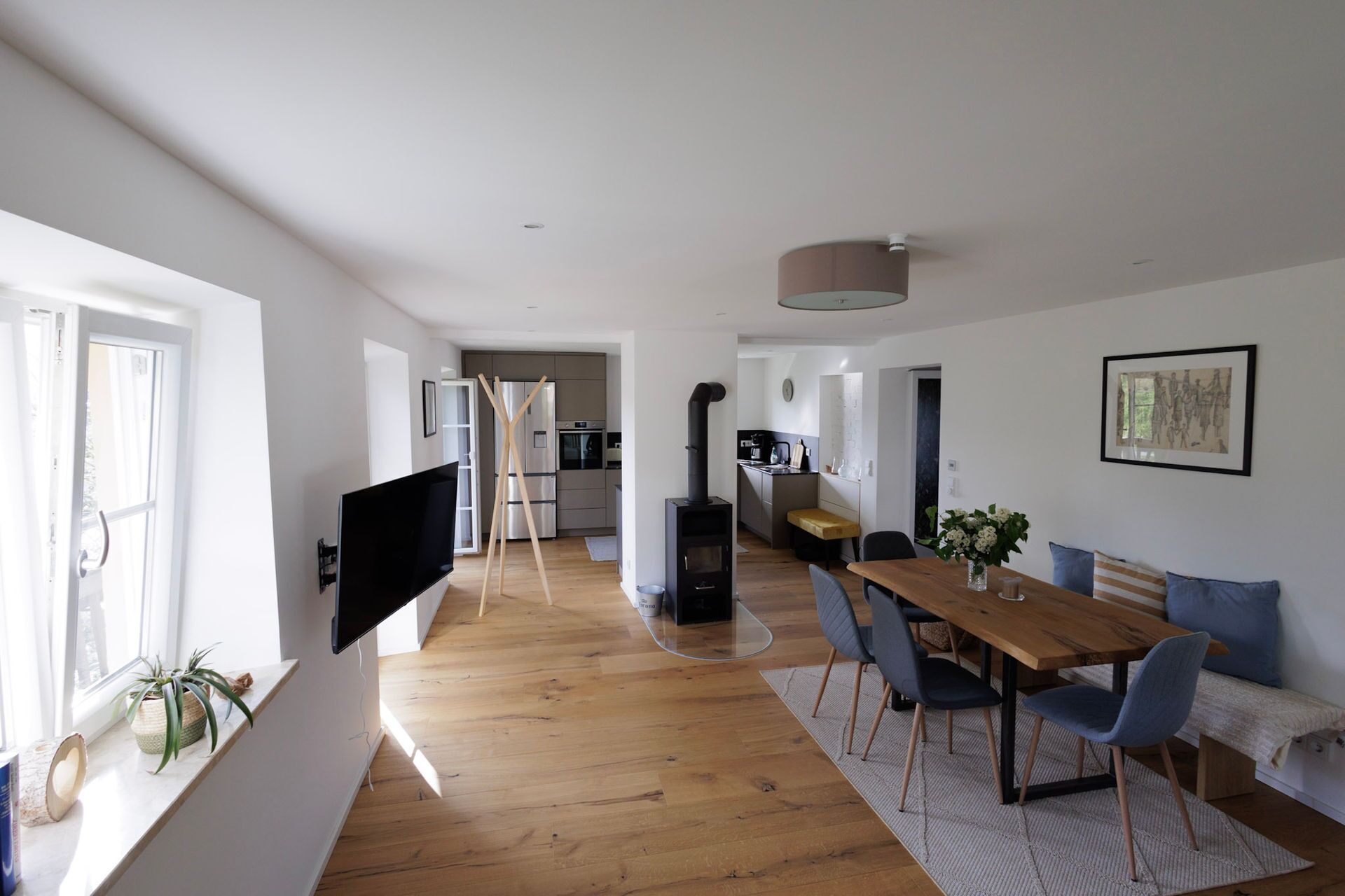 Ein modernes Wohnzimmer mit einem Holzesstisch, blauen Stühlen, einem an der Wand montierten Fernseher, einer Küchenzeile und einem großen Fenster.