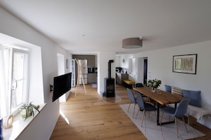 A modern living space with a wooden dining table, blue chairs, a TV mounted on the wall, a kitchen area, and a large window.