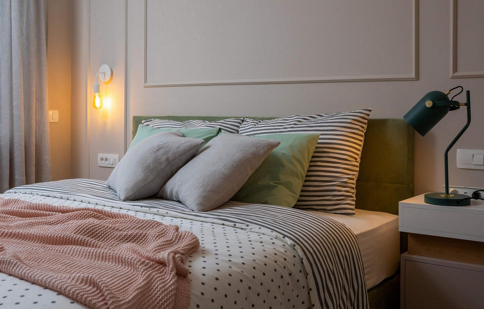 A neatly made bed with striped bedding, a green headboard, and two pillows. There is a bedside lamp and a power outlet on the nightstand.