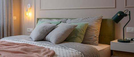 A neatly made bed with striped bedding, a green headboard, and two pillows. There is a bedside lamp and a power outlet on the nightstand.