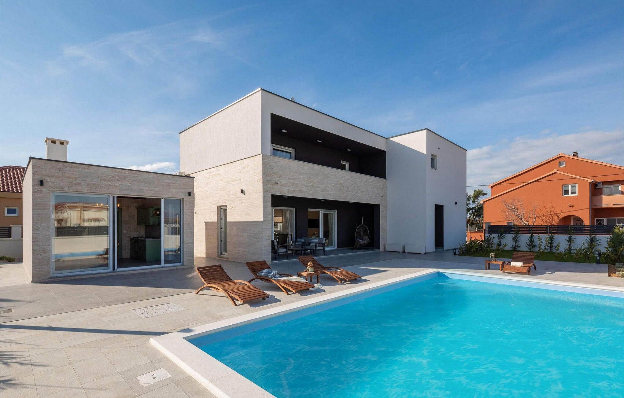 A modern two-story house with a swimming pool, outdoor seating, and a large glass door entrance.