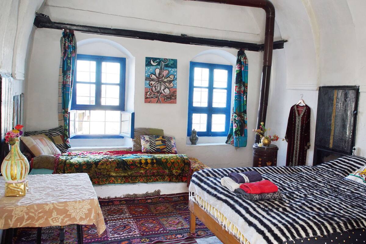 Une chambre avec deux lits, une table sur laquelle se trouve un vase, et une fenêtre avec des volets bleus.