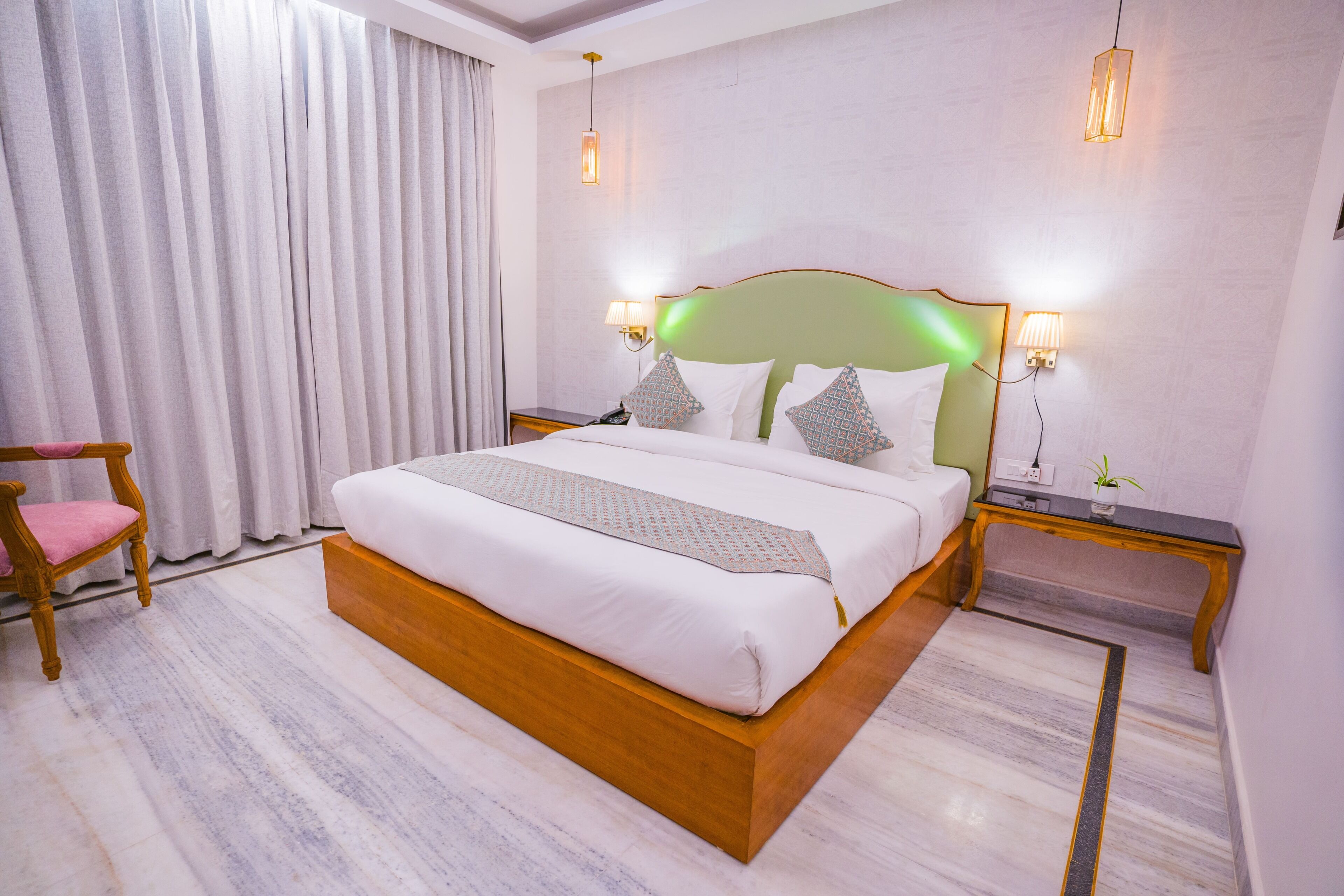 Una camera d'albergo con un letto grande, una testiera in legno, due comodini, una sedia e un'ampia finestra con tende.