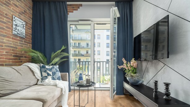 A modern living room with a sofa, a small table with a tray, a TV mounted on the wall, and a balcony with a view of buildings. - MARGI Soho Apartament (Szczecin)