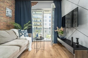 Een moderne woonkamer met een bank, een klein tafeltje met dienblad, een aan de muur bevestigde televisie en een balkon met uitzicht op gebouwen.
