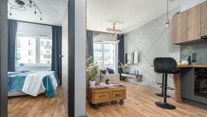 A modern living room with a sofa, a coffee table, and a dining area with a chair and a table. There is a window with curtains, a plant, and a chandelier. - MARGI Soho Apartament (Szczecin)