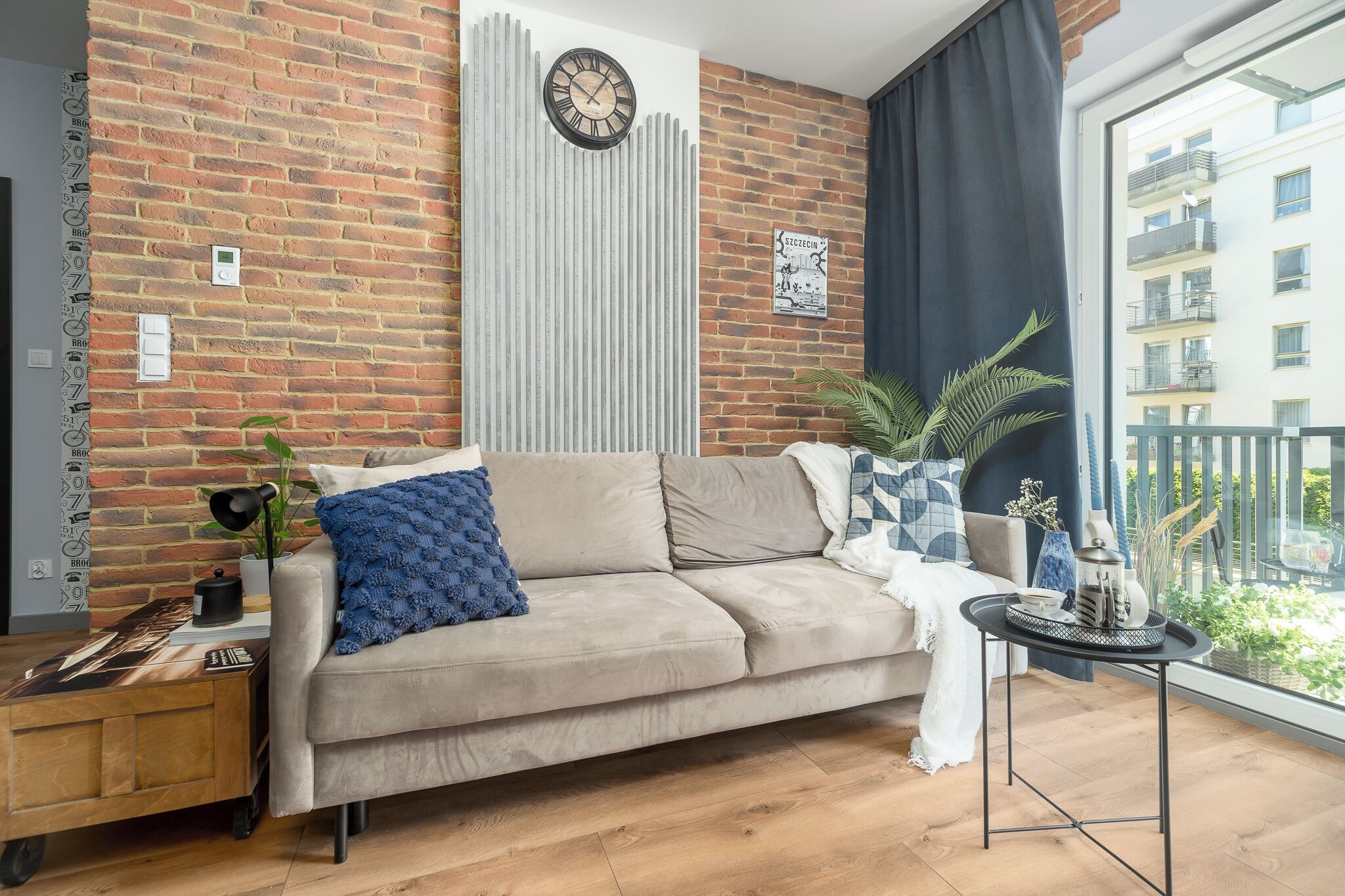 A living room with a brick wall, a sofa, a side table, and a view of a building through a window.