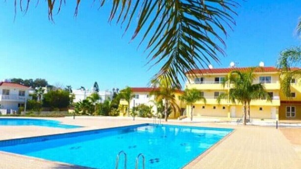 A swimming pool with clear blue water, surrounded by palm trees and a building with a red roof. - 2 bedroom with private garden, walk to Nissi Beach
 (Ayia Napa)