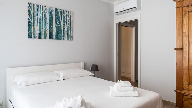 A neatly made bed with white linens and towels, a wall-mounted painting, and a wooden wardrobe.