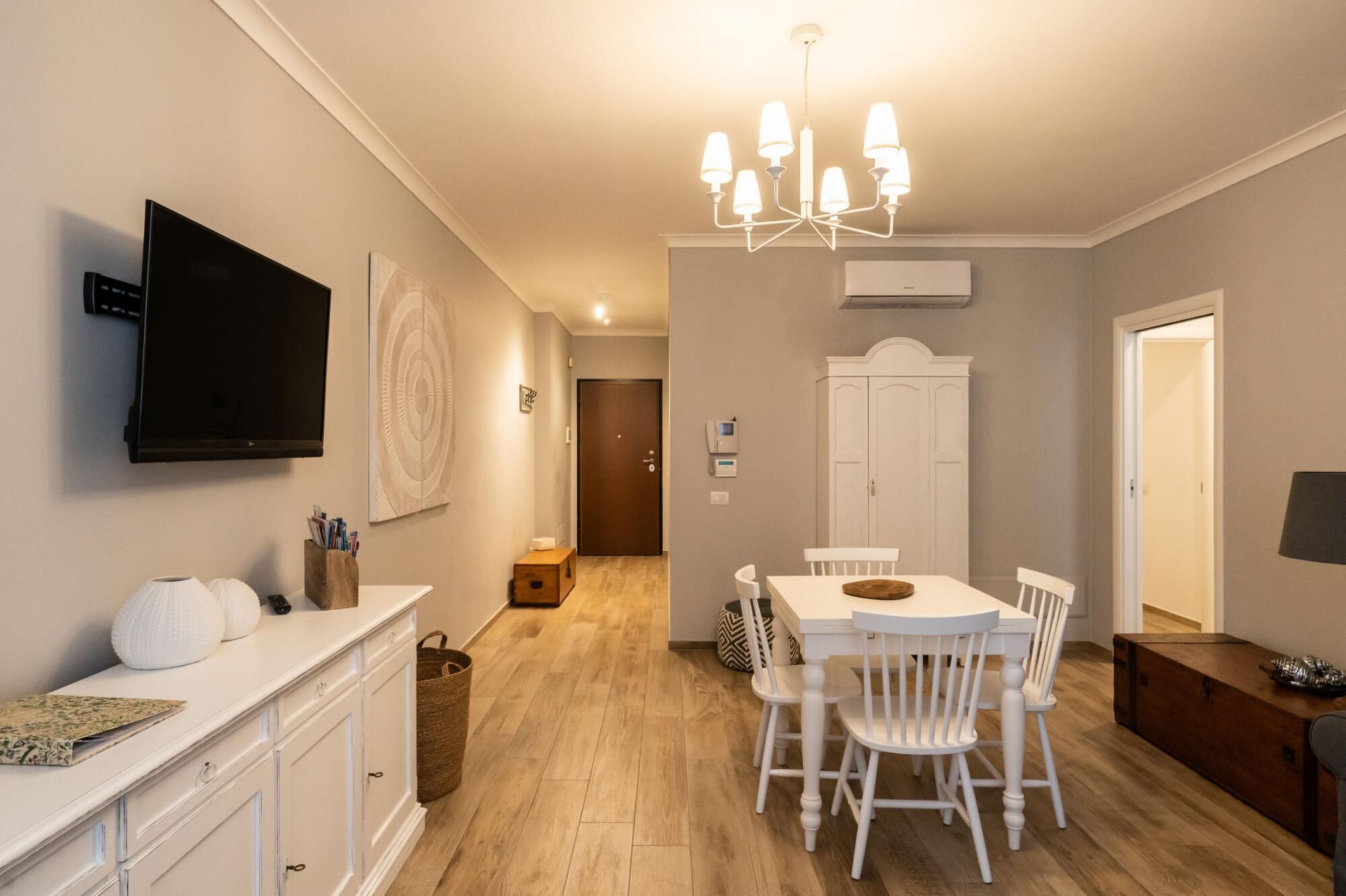 A modern kitchen with a dining area, white cabinetry, a flat-screen TV, and a chandelier.