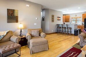 A spacious living room with a sofa, ottoman, and a lamp. A kitchen with a bar and stools is visible in the background.