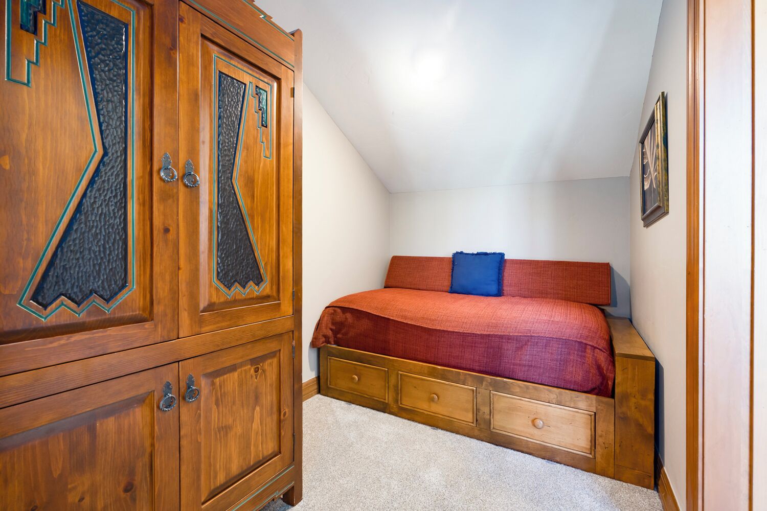 A bedroom with a wooden wardrobe, a bed with a red blanket, and a small bedside table.