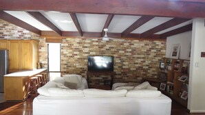A living room with a sofa, a television on a brick wall, a kitchen area with wooden cabinets, and a ceiling fan. - Beautiful house, located in a quiet area of town with its own private pool (South West Rocks)