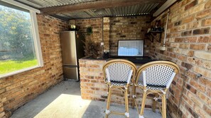 A compact kitchen with a brick wall, a stainless steel refrigerator, a small bar area, and two wicker chairs.