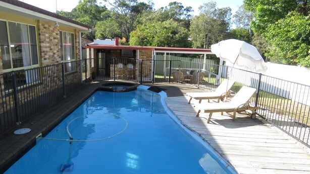 A swimming pool with a wooden deck and lounge chairs, surrounded by a fence and trees. - Beautiful house, located in a quiet area of town with its own private pool (South West Rocks)
