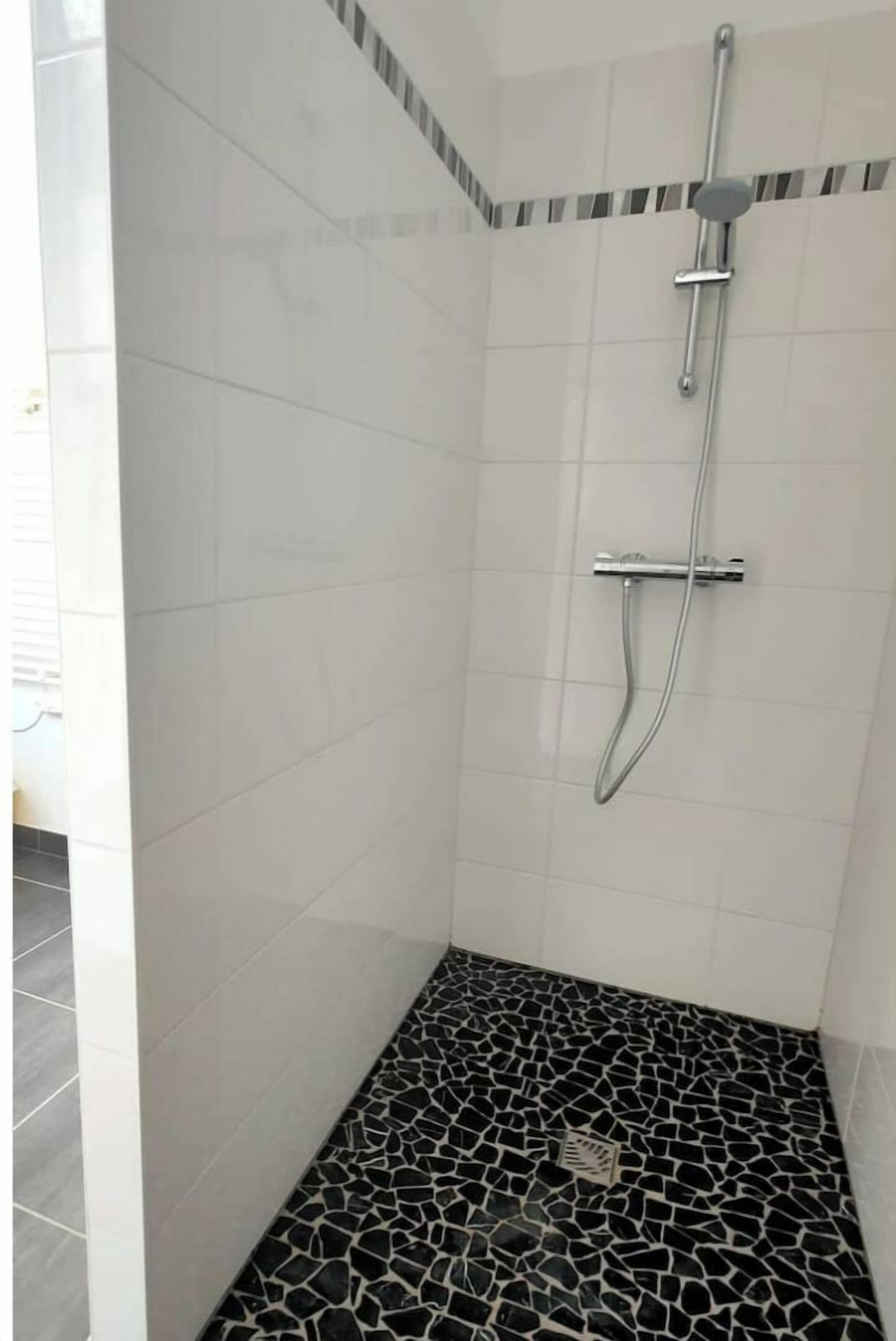 A shower with a handheld showerhead and a wall-mounted faucet, tiled walls, and a floor with a black and white mosaic pattern.