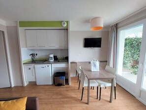 A compact kitchen with white cabinets, a coffee maker, and a dishwasher. There's a dining table with chairs, a TV mounted on the wall, and a window with curtains.