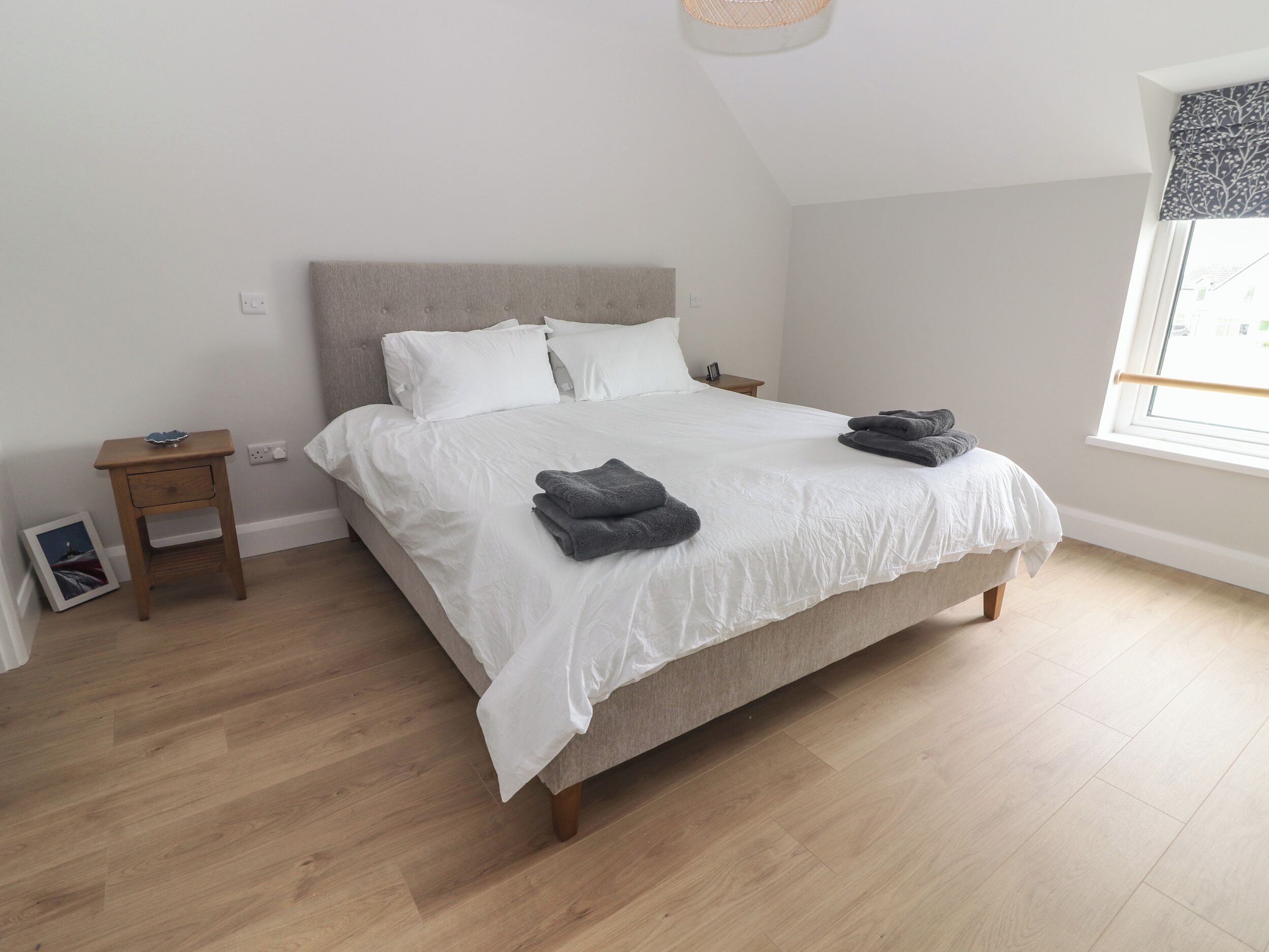 A neatly made bed with white linens and folded towels, a small wooden nightstand with a lamp, and a window with a view of the outside.