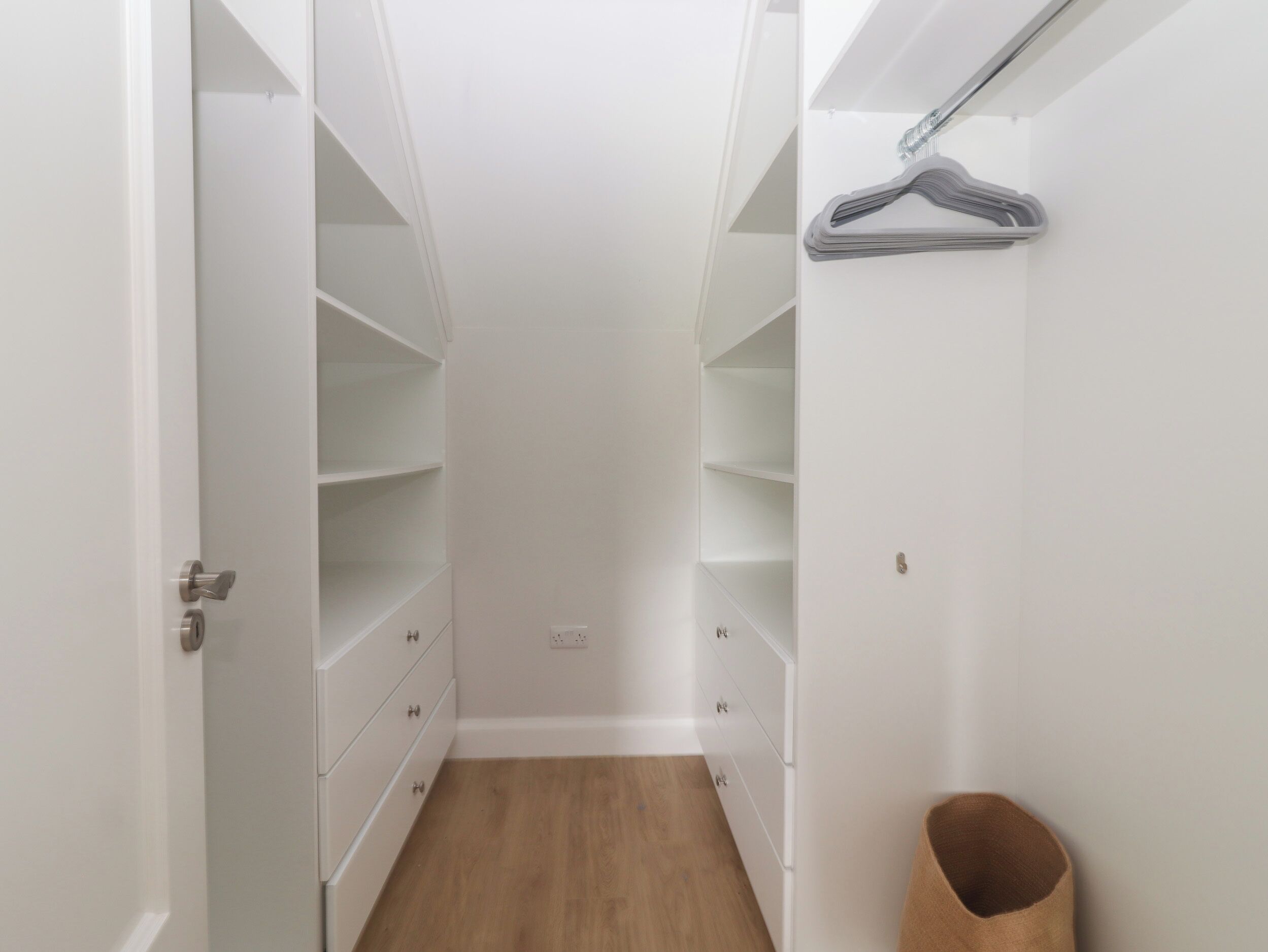 Un petit dressing étroit avec de nombreuses étagères et des tiroirs, un plancher en bois et une tringle à vêtements fixée au mur de droite.