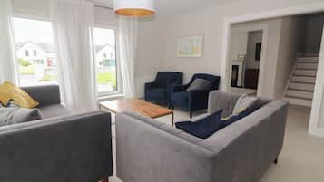 Un salón moderno con sofás grises, una mesa de centro de madera y sillones azules. Hay un ventanal con cortinas blancas, una lámpara colgante y un cuadro en la pared.